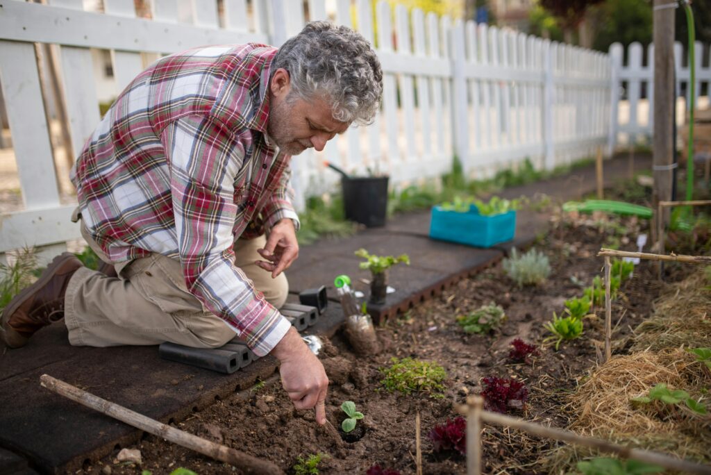 An elderly man kneels to plant in a vibrant garden, surrounded by tools and greenery.